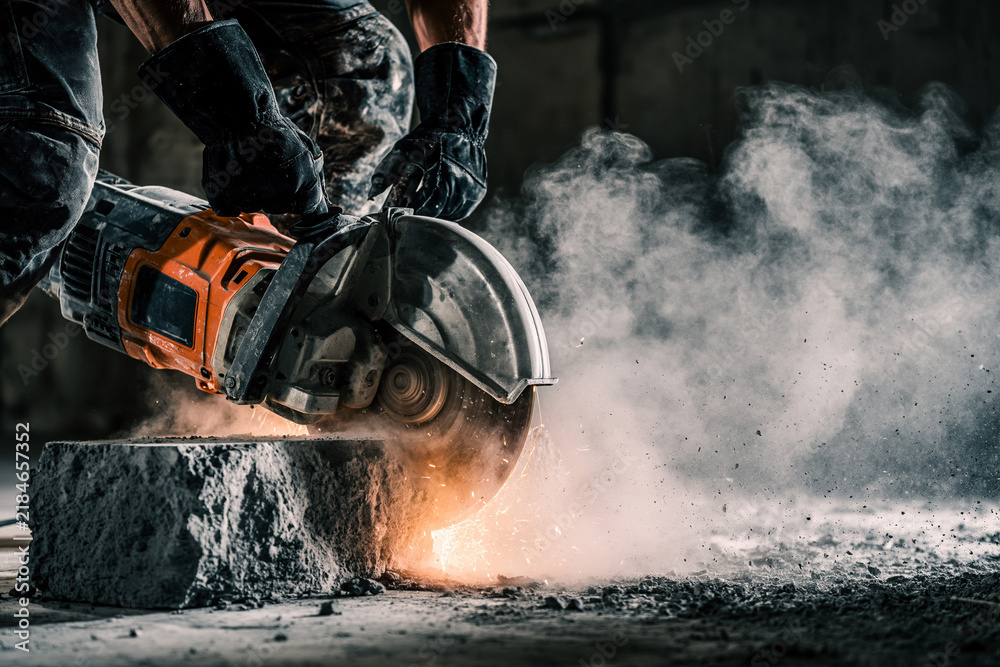 Obraz premium A worker in dark, soiled work and black gloves uses a handheld orange and grey circular concrete saw.