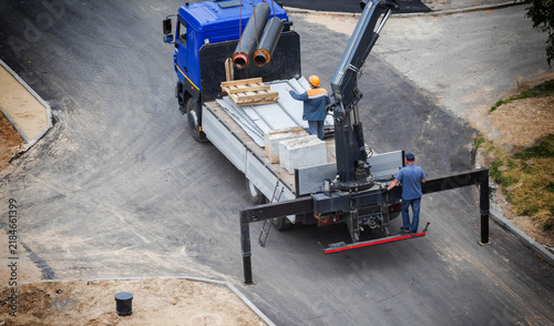 Blue construction truck equipped with hydraulic manipulator crane system unloads large insulated pipes for infrastructure project. Two workers assist with heavy material handling on road construction