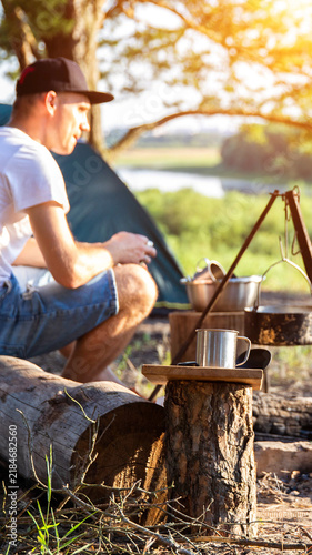 Adult male camper crouches near campfire setup with cooking pots and metal mug outdoors at sunny campsite by river, tent and lush foliage in background, vertical photo