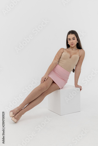 A beautiful young woman in a beige bodysuit and a pink skirt, wearing mules, sits on a white cube against a white background.