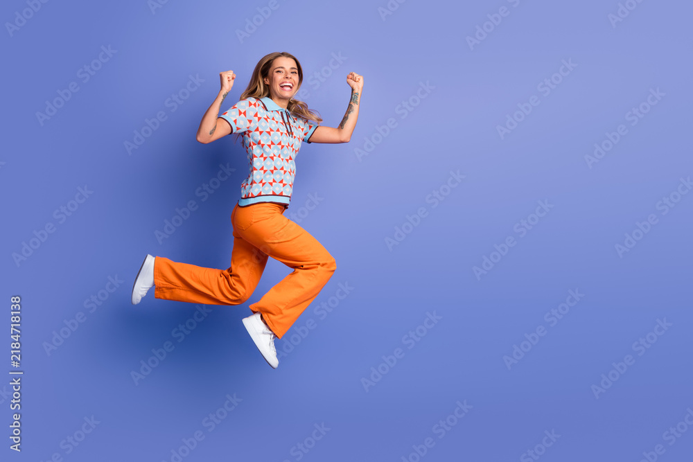 Fototapeta premium Happy young woman leaps in mid air against a blue background wearing orange pants and a patterned polo top