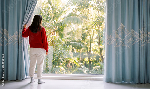 Morning in the tropics. A girl in a red sweater opens the blue curtains and looks through the panoramic window at a lush tropical garden on a sunny day