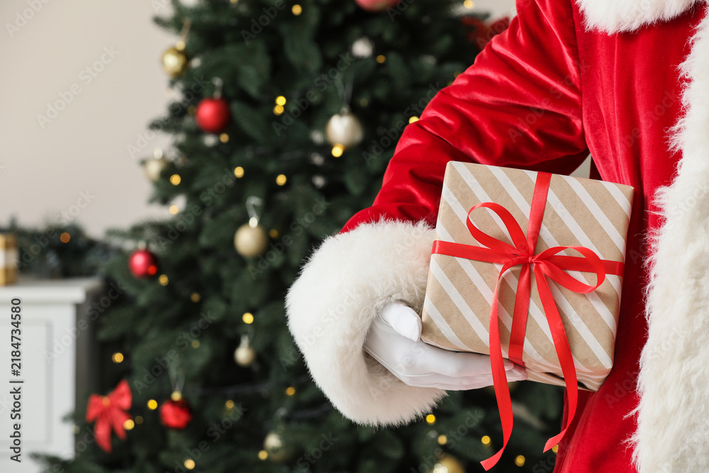 Obraz premium Christmas gift giving and holiday celebration, Santa Claus holding a wrapped present with a red ribbon in front of a decorated pine tree at home
