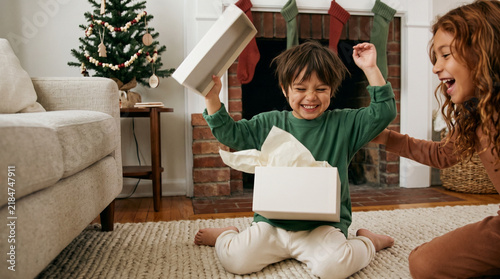 Happy kid unwrapping a holiday gift beside a parent indoors, cheerful seasonal moment with excitement and Christmas magic  