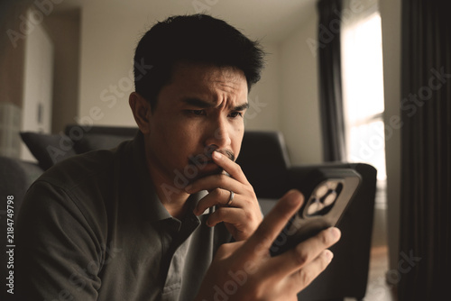 Man Wearing a Concerned Expression While Holding and Reading His Phone Near a Window
