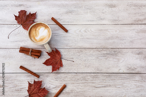 Ceramic cup of coffee with milk, red maple leaves and cinnamon on wooden table. Autumn hot drink, mug of latte on white wooden background. Top view