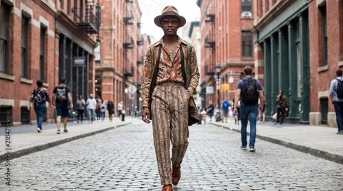 Stylish African American man in patterned suit and hat walks confidently down a cobblestone street surrounded by historic brick buildings and pedestrians in a vibrant urban setting