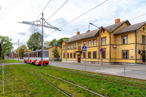 Red and white tram on green grass tracks passing by traditional yellow wooden houses under an overcast sky in the Kalamaja district of Tallinn, Estonia.
