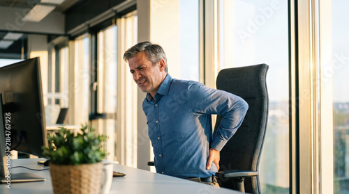 Middle aged man gripping lower back in modern office with pain and discomfort during workday