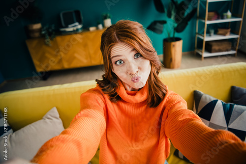 Obraz na plátně Playful young woman taking a selfie on a yellow couch in a colorful living room
