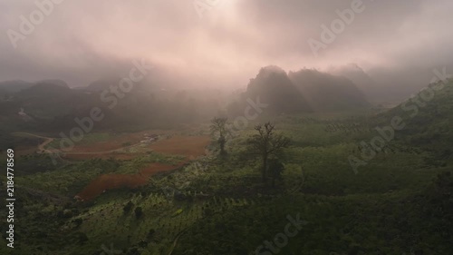 Atmospheric aerial view of mist-covered mountains, rural farmland, terraced fields, and soft golden light filtering through clouds in a peaceful valley.