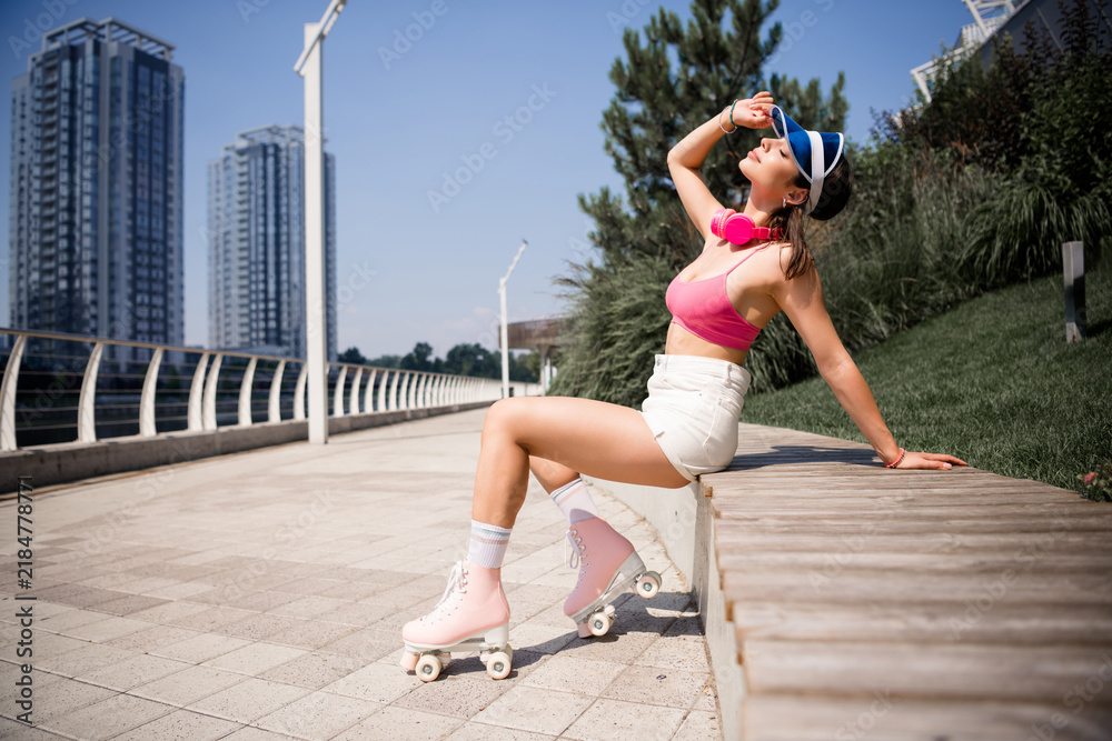Fototapeta premium Young woman roller skating in pink outfit relaxing on an urban waterfront bench on a sunny summer day