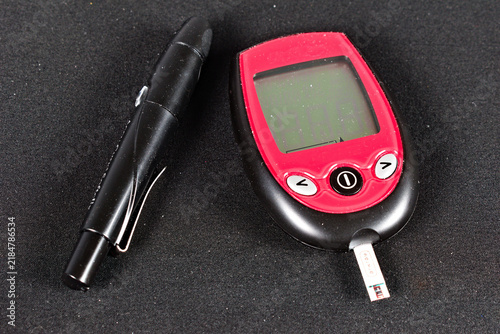 фотографія A red and black glucose meter with a test strip inserted sits next to a black lancet pen on a textured dark surface