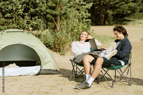 Young adult Caucasian couple relaxing by tent outdoors while working on laptop. Suitable for digital nomad lifestyle  remote work campaigns  and modern outdoor leisure marketing