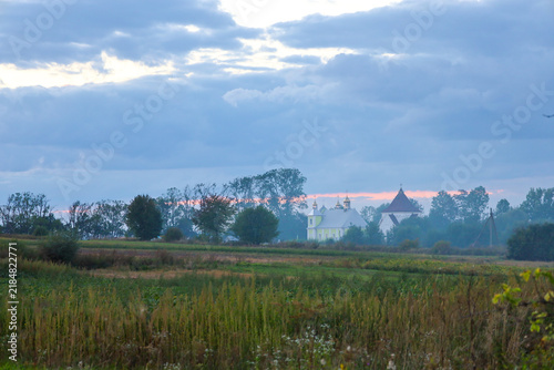 Cuadro en lienzo A picturesque rural church and majestic trees rise behind a green field under a cloudy evening sky