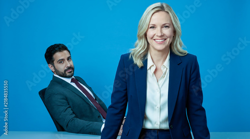 Portrait of Smiling Businesswoman Standing with Colleague Seated Against Blue Background