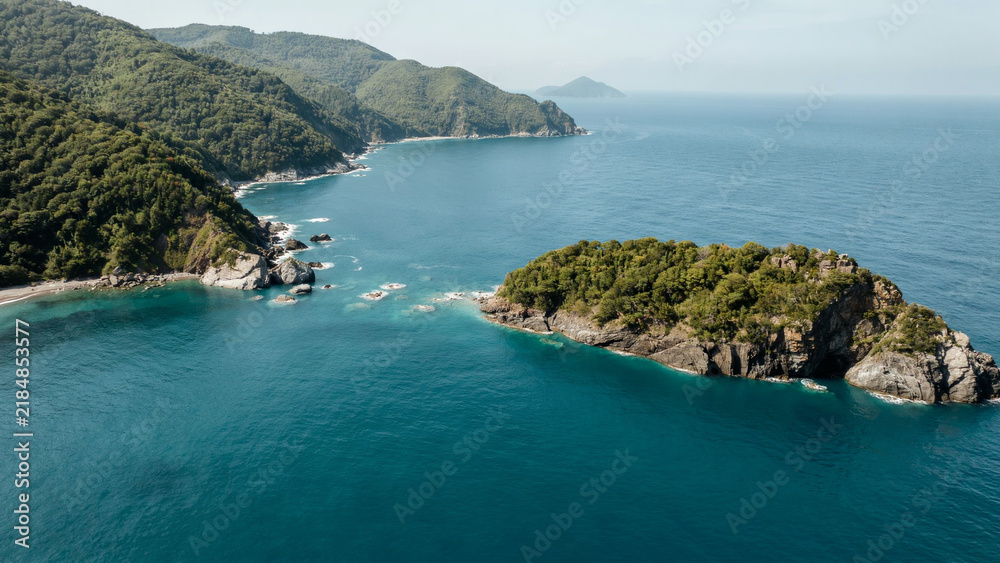Obraz premium Aerial view of lush green mountains meeting turquoise ocean waters near rocky coastline.
