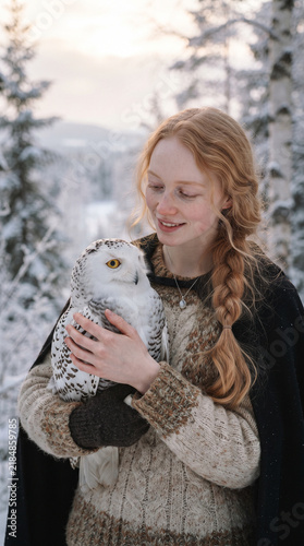 Young fair-haired woman gently cradling a speckled owl outdoors in a snowy forest setting during golden hour light