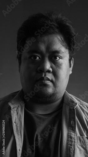 Black and white portrait of a young Asian male staring directly at the camera with a neutral, intense expression. The subject has short, slightly unkempt dark hair and a short beard,