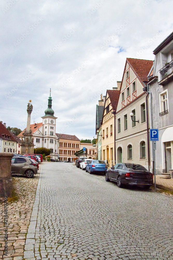 Obraz premium Eine gepflasterte Straße führt durch das historische Zentrum von Loket zu einem Uhrturm und einer barocken Säule; entlang der Häuser stehen Fahrzeuge.