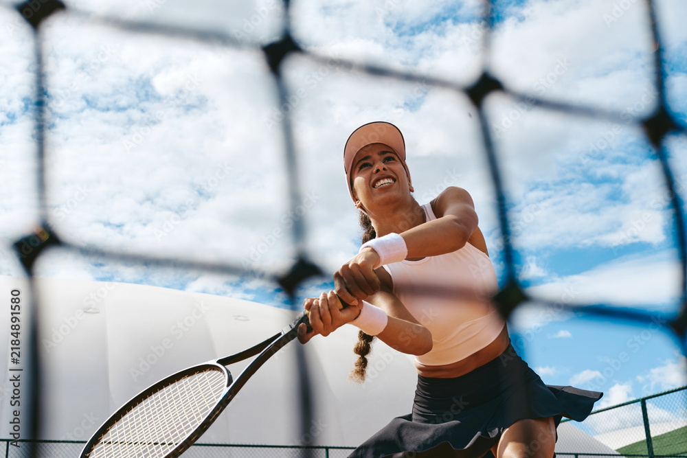 Fototapeta premium Low angle view of multiethnic young adult woman playing tennis, hitting two handed backhand on court. Image ideal for sports advertising and fitness promotion