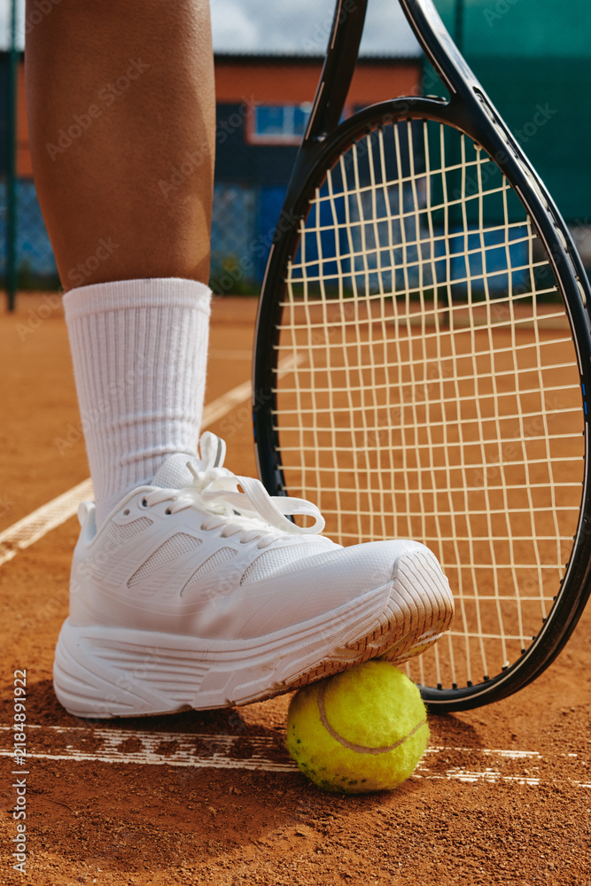 Fototapeta premium Vertical shot of woman foot stepping on tennis ball next to racket on clay court during training match. Image suitable for sports marketing and active lifestyle campaigns