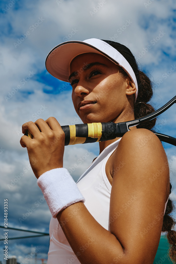 Fototapeta premium Vertical shot of young multiethnic woman holding racket on outdoor tennis court before match. Imagery suitable for sports branding, athletic apparel campaigns and fitness marketing