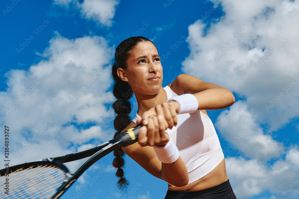 Fototapeta premium Low angle view of young adult multiethnic woman playing tennis, hitting backhand stroke outdoors. Visual concept for fitness campaigns, sports advertising, athletic training