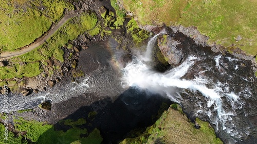 Aerial view of a powerful waterfall cascading over rocky cliffs into a pool with a faint rainbow in the mist, surrounded by vibrant green mossy terrain in Iceland.