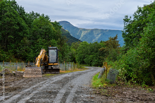 Quadro su tela Heavy machinery stands idle on a rugged road lined with trees as clouds gather over majestic mountain ranges