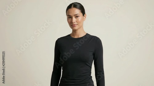 Confident young Asian woman wearing a black long-sleeve athletic top posing against a neutral beige background in a minimalist studio setting.