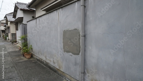 Plakat w ramie 灰色の塀に残る四角い補修跡と細いひび割れや雨樋が目立ち足元に植木鉢が並ぶ日本の住宅路地