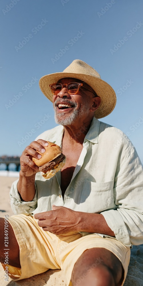 Fototapeta premium Smiling man in straw hat eating a burger on the beach under a clear blue sky for summer lifestyle and outdoor picnic banner design