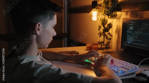 A teenage boy typing code on an RGB mechanical keyboard while working on a laptop at a desk in a dark room illuminated by warm lighting.
