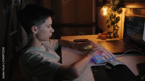Teenage boy typing on an RGB keyboard and coding on a laptop at a desk in a dimly lit room at night.
