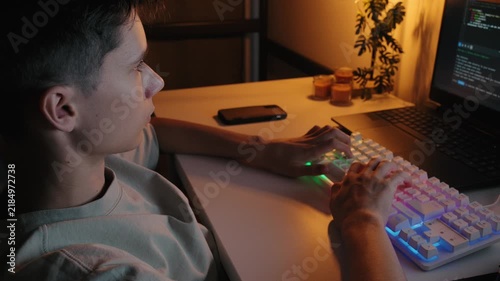 Focused teenage boy coding on laptop and typing on RGB backlit keyboard at desk in dark room.