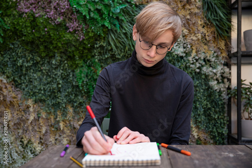 A young man wearing glasses drawing in notebook with pencil