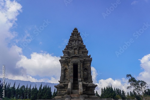 Ancient stone temple tower rises against a clear blue sky with billowing clouds, framed by distant mountains and evergreen trees, suggesting history, spirituality, travel and architectural heritage