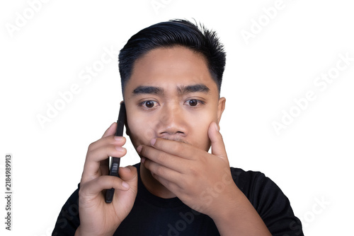 фотографія Shocked Indonesian man wearing a black t-shirt talking on a mobile phone while covering his mouth with his hand and looking wide-eyed against a white background