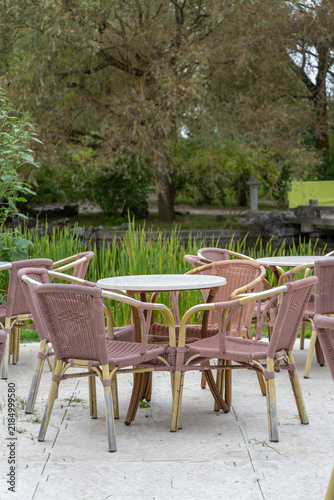Cuadro en lienzo Empty pink wicker chairs surround round white tables on a stone patio, with tall reeds and a pond visible beyond a willow tree