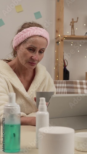 Mature woman wearing a facial sheet mask drinks tea and uses a digital tablet on a sofa at home, enjoying a relaxing beauty routine.