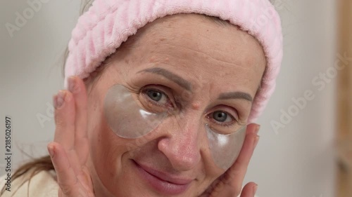 Portrait of a senior woman applying hydrogel eye patches while caring for aging facial skin during her beauty routine at home.