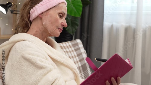 Middle aged woman in a bathrobe and facial sheet mask writes and plans in a personal journal while sitting on a sofa at home