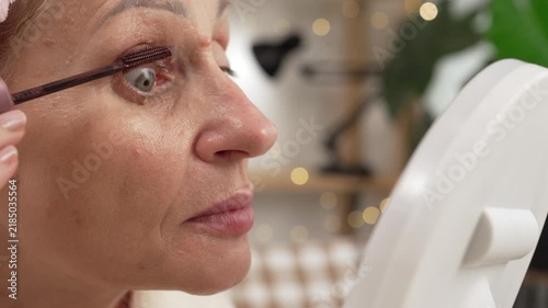 Elderly woman applying mascara to her eyelashes while doing evening makeup at home.