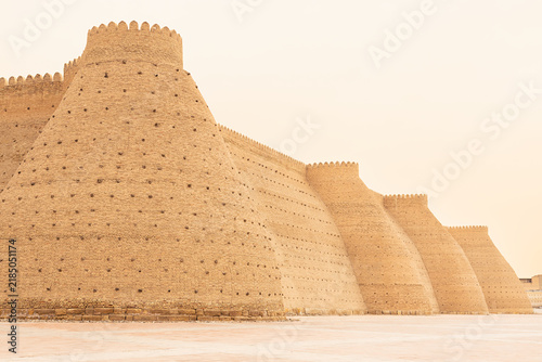 Ark fortress massive brick walls with repeating towers in Bukhara Uzbekistan. Ancient citadel exterior with crenellated parapets under hazy sky. Ideal for travel publications and heritage content.