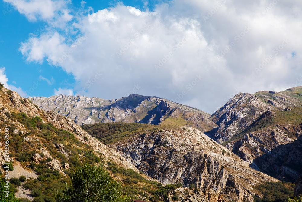 Fototapeta premium Rugged mountains stretch beneath a vibrant blue sky. Clouds drift above jagged peaks and rocky ridges. Earth tones dominate the landscape, with green accents. Sunlight highlights the textured stone