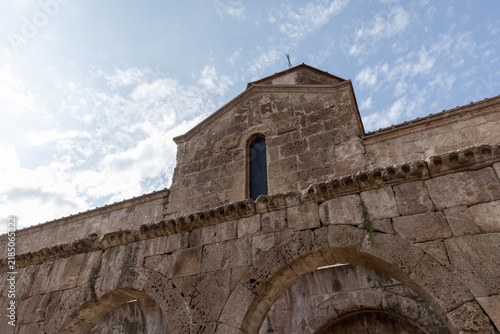 Quadro su tela Historic stone church facade featuring rounded arches, a narrow arched window and weathered masonry against a bright blue sky