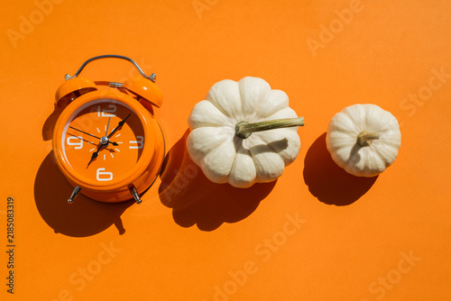 Orange alarm clock with three white decorative pumpkins on two tone beige and orange background for autumn season and daylight saving time concept