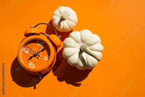 Orange alarm clock with three white decorative pumpkins on two tone beige and orange background for autumn season and daylight saving time concept