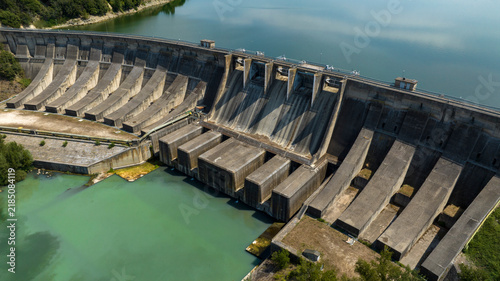 Obraz na plátně Aerial close-up view of a massive concrete hydroelectric dam wall, showing its intricate spillway structures and holding back a large body of water under a bright sky with reflections on the surface